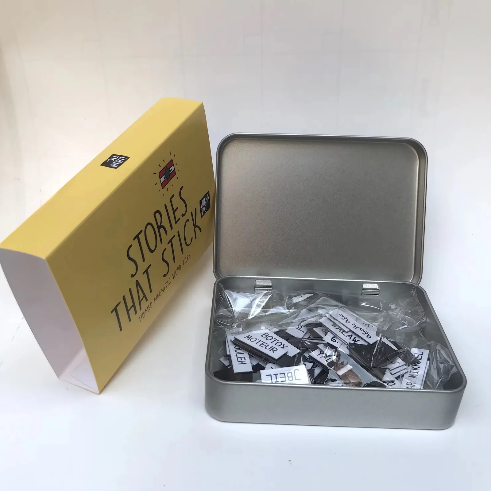 A tin of magnetic word tiles next to its printed yellow sleeve box, showing set packaging for a gift-level product